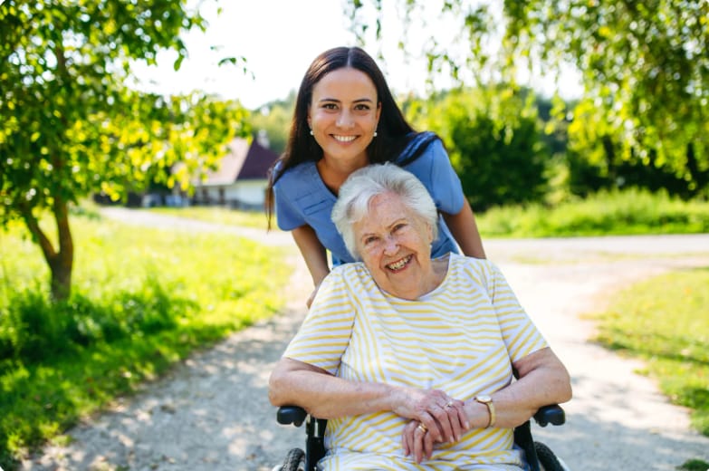 Betreuungskraft von Helfen mit Herz schiebt eine lachende Seniorin im Rollstuhl über einen sonnigen Weg