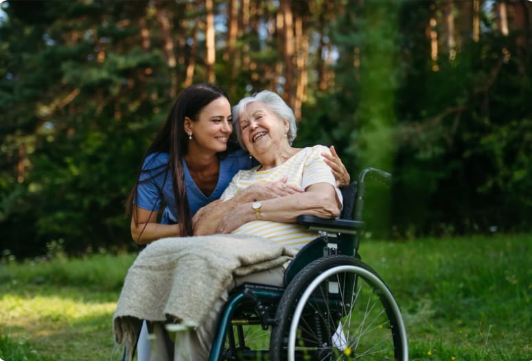 Betreuungskraft umarmt eine Seniorin im Rollstuhl – beide lachen herzlich